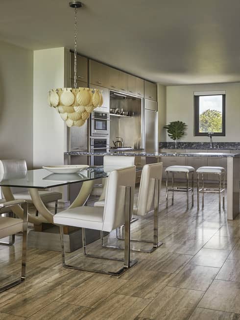Luxury dining area with a glass table, white leather chairs, decorative circular mirror, shell chandelier and adjacent kitchen with stainless steel appliances