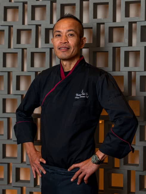 Man with short dark hair wears a black chef's jacket with red trim and stands with arms akimbo in front of a wall comprised of hollow grey squares