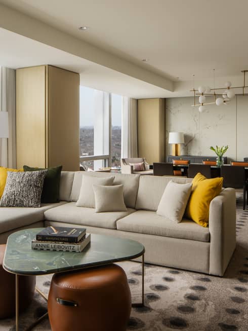 Spacious suite living area with a large sectional sofa, yellow and gray pillows, marble coffee table, and a dining area with elegant décor and city views