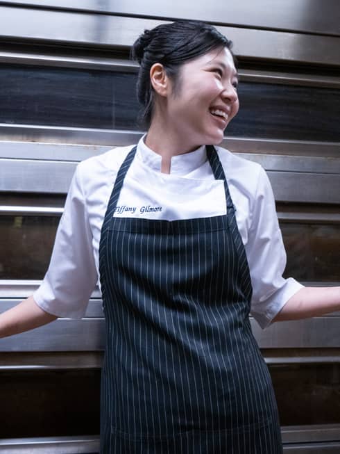 A chef in a white shirt and a blue apron stands in a kitchen setting