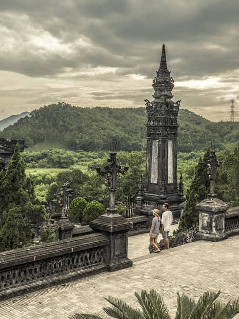 Man and woman walk down steps of historic Imperial City of Hue with stone towers, rolling hills