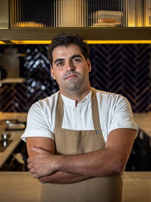 Chef Rodolfo Lavrador in white short-sleeve shirt and beige apron with arms crossed, standing in front of open kitchen