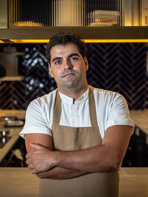 Chef Rodolfo Lavrador in white short-sleeve shirt and beige apron with arms crossed, standing in front of open kitchen