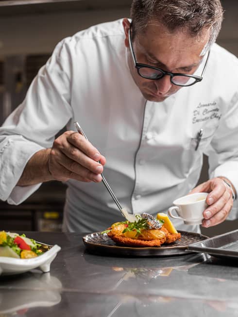 A chef lightly pours a sauce onto a plate of chicken and herbs.
