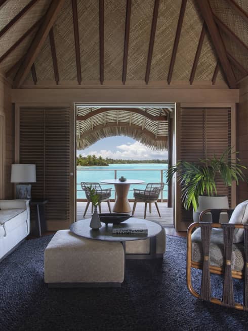 Overwater bungalow living area with neutral furniture, wood beams, tropical plants and a patio with a view of the turquoise lagoon.