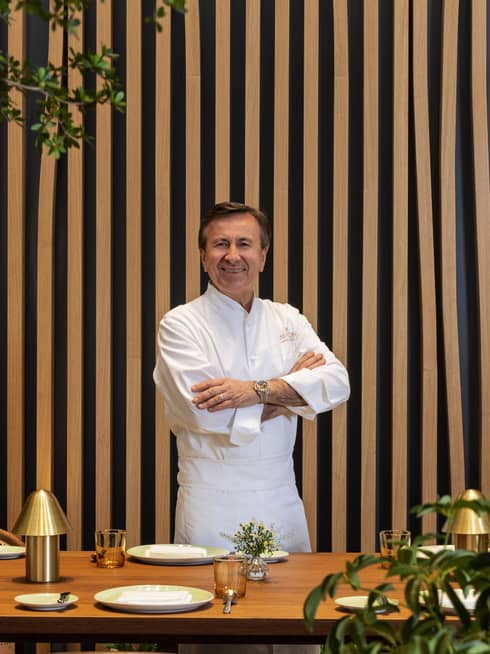 A chef in a white shirt and trousers stands in front of a pin-striped wall surrounded by foliage.