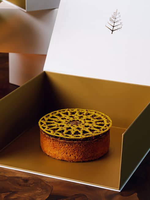 Round dessert with a lattice topper sits in the middle of an open gold box with a white lid that has the Four Seasons tree logo in the centre shows