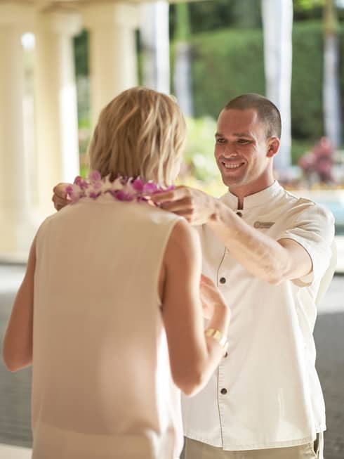 Hotel staff member smiling while placing a purple flower lei on a guest during arrival