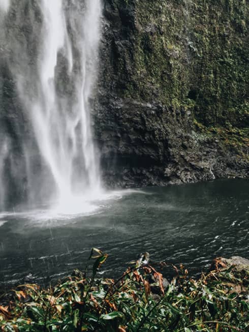 Back of woman hiking over black rocks under tall waterfall, small pond