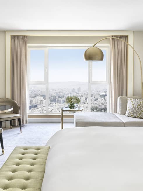 Hotel room with white king bed, light green cushioned bench, two taupe chairs, chaise, wall of windows