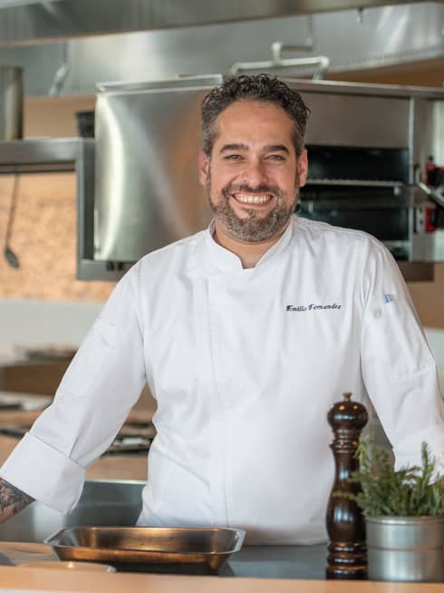 Executive Chef Emilio Fernandez in white chef's coat, standing in restaurant kitchen, smiling