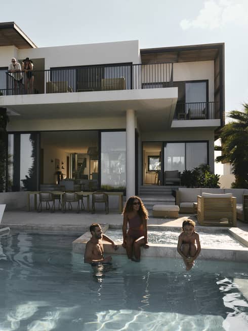 A woman, man and young child sit on edge of private pool at luxury residence in tropical setting