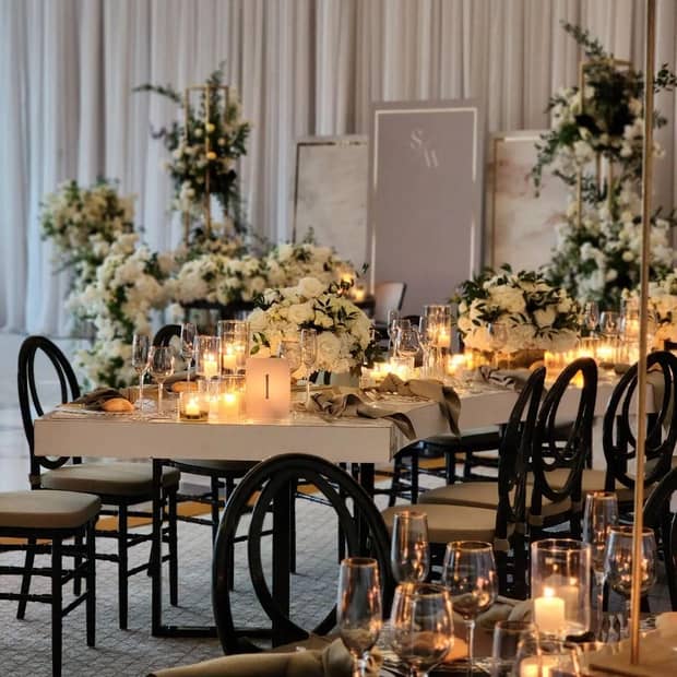 A candlelit wedding banquet table decorated with greenery and white floral arrangements of various sizes, linen napkins, wine and champagne glasses