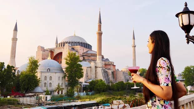 Woman holding red cocktail stands on balcony looking out at Hagia Sophia museum in Istanbul