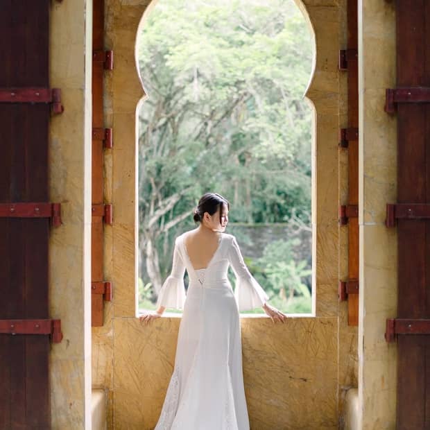 ,A woman in a white wedding dress stands in front of a large window overlooking green foliage