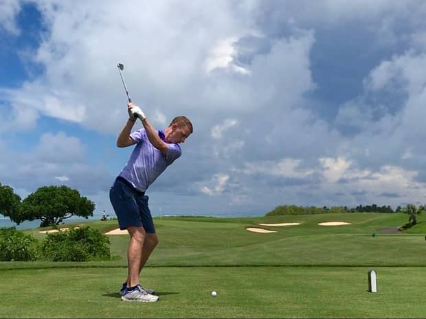 Man swings golf club on rolling green course under blue sky, white clouds