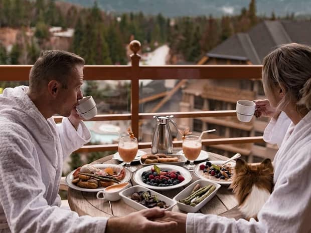 Couple wearing bathrobes, small dog on lap, sip coffee at private balcony breakfast table