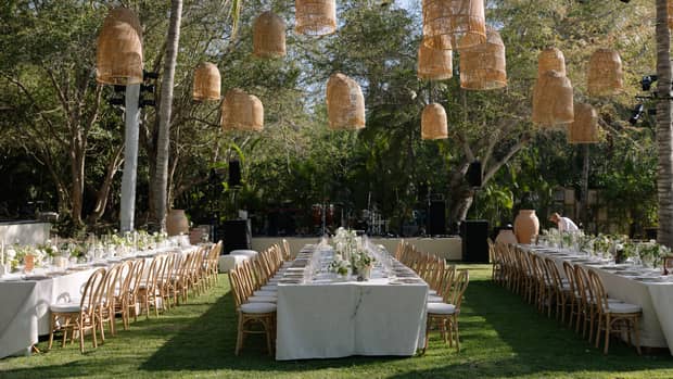 Long dining tables with seating in an outdoor event space surrounded by trees. Tables are set for an event, complete with floral arrangements and outdoor pendant lighting.