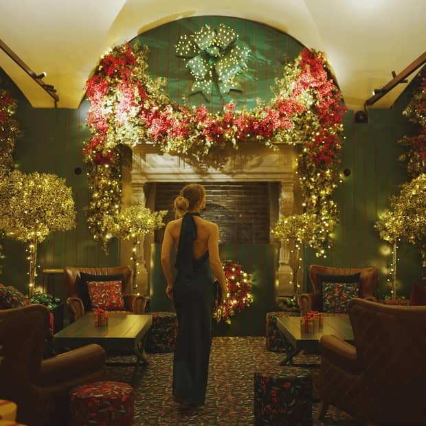A woman in a long gown walks through a lounge with leather chairs and wooden tables, towards a fireplace adorned with red and gold festive garland with twinkling lights