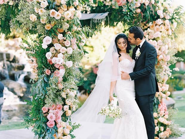 Groom kisses bride under wedding altar covered with pink roses on outdoor patio