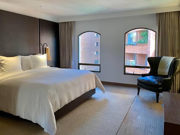 Modern hotel room with bed against panel wall, velvet wingback armchair by two sunny windows