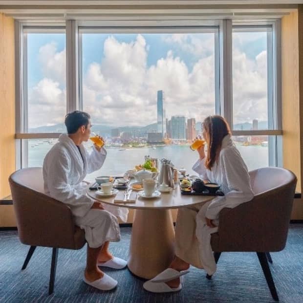A couple dressed in white bath robes and slippers dine in front of a large window overlooking a city skyline