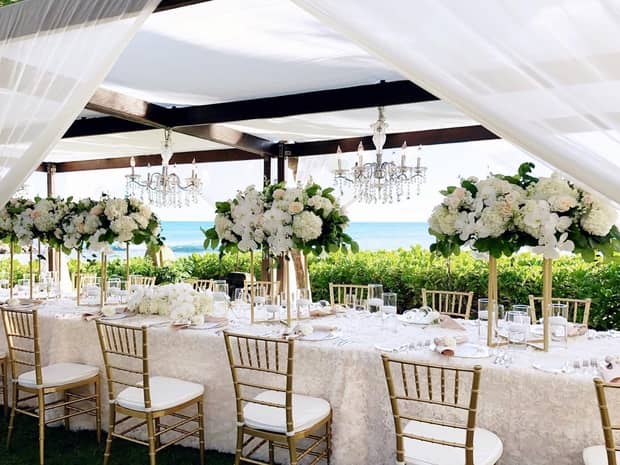 A lavish wedding dining table set in serene outdoors, adorned with white flowers, crystal chandeliers, overlooking the sea