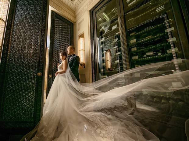 A bride in a voluminous gown and groom in a suit stand in an elegant setting with a luxurious French fine dining backdrop, intricate designs, and lighting.