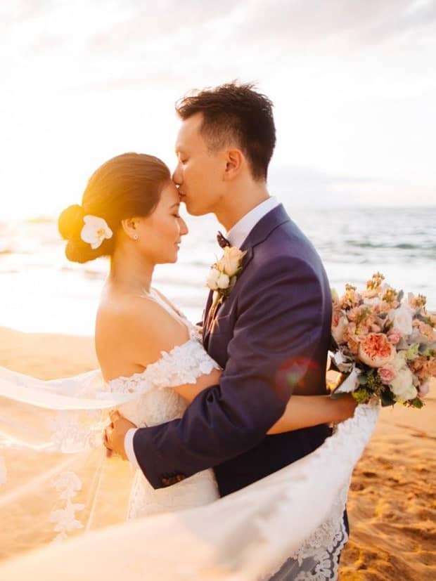 Groom kisses bride's forehead on beach at sunset