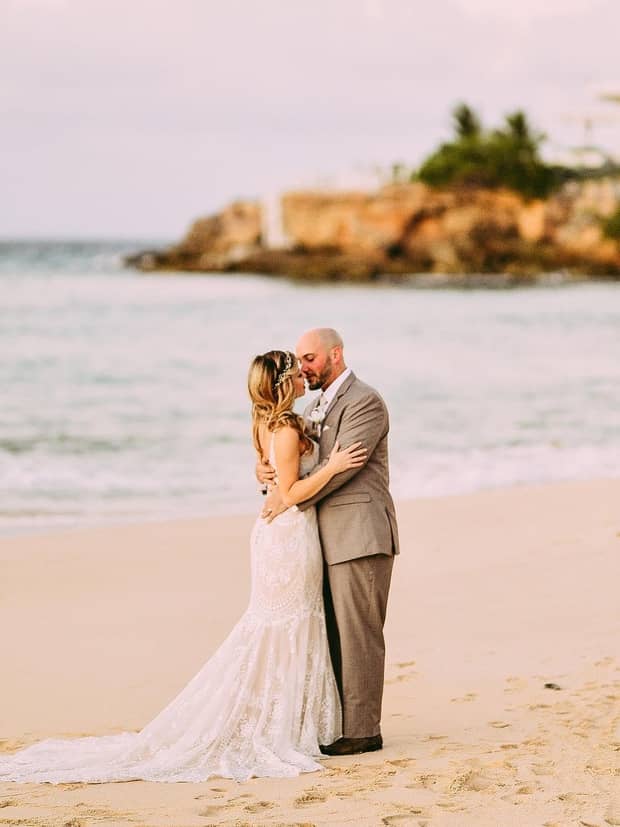 Couple embracing on a sandy beach at sunset, dressed in wedding attire.