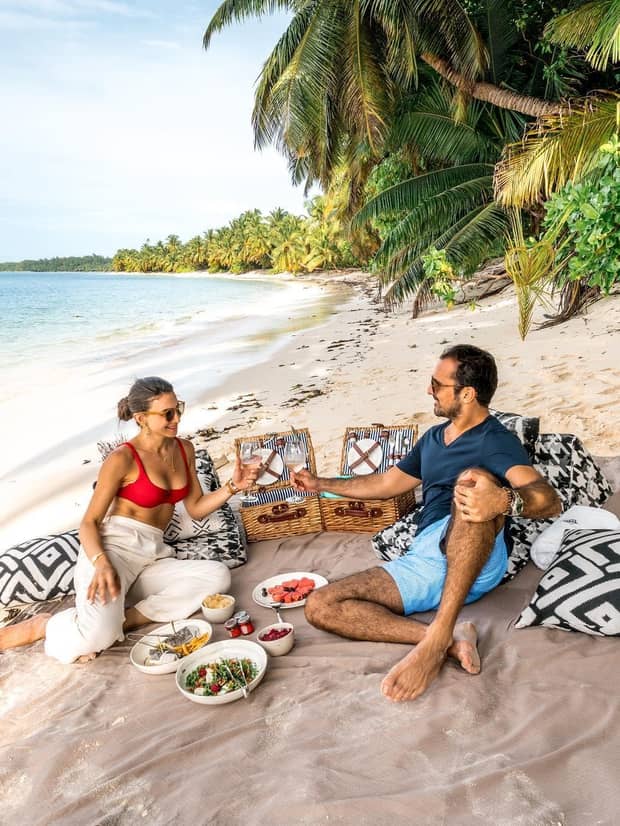 Couple lean back on wicker baskets, enjoy picnic lunch on white sand beach