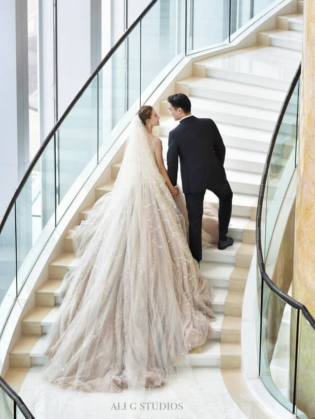 A bride in a flowing gown and a groom in a black suit hold hands on a spiral staircase. The bride's veil cascades down the steps as they look at each other.