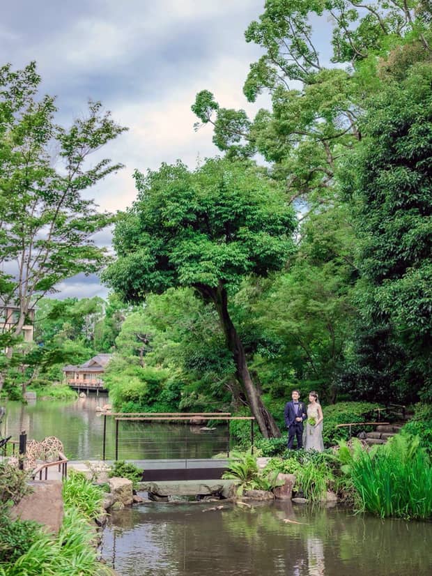Bride and groom stand before small bridge and pond surrounded by trees