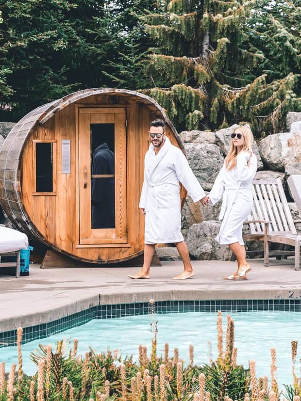 Couple wearing bathrobes walk on outdoor swimming pool deck past small steam rooms