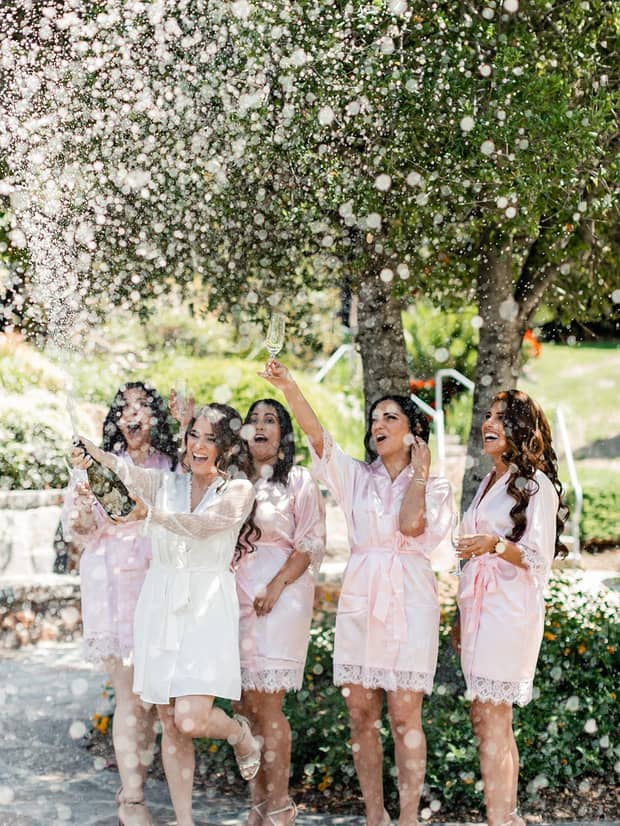 Bride and bridal party wearing matching robes laugh, pop Champagne in outdoor space