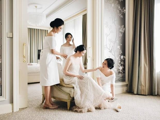 Smiling bridesmaids help bride with her shoes as she sits in armchair