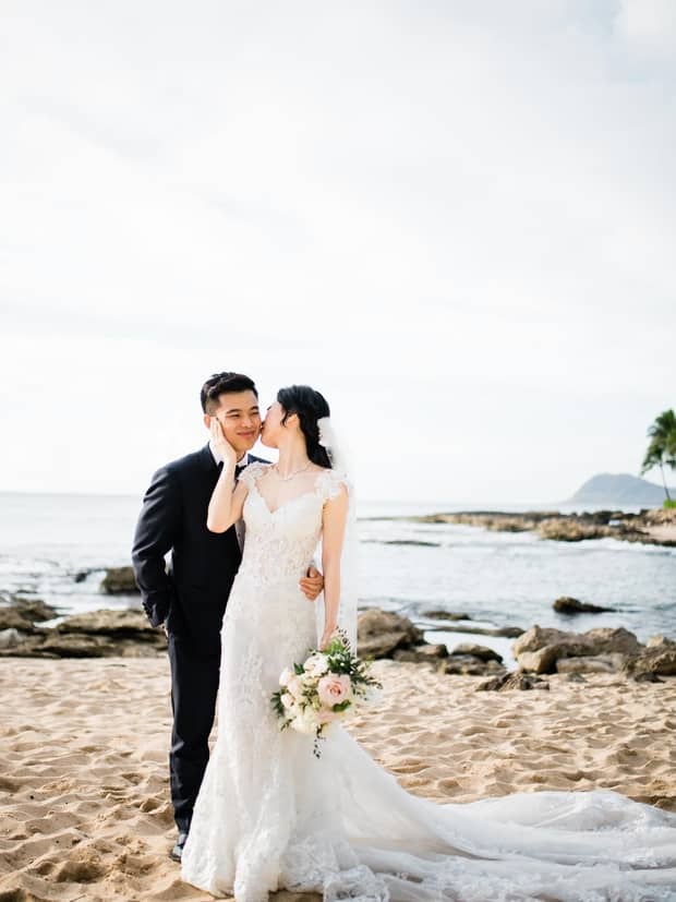 A romantic wedding photo of a bride kissing groom's cheek, set against a stunning backdrop of white sand beach and clear blue sky