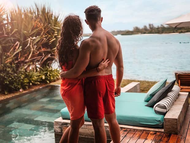 Back of couple wearing swimsuits standing by patio swimming pool and lounge chair