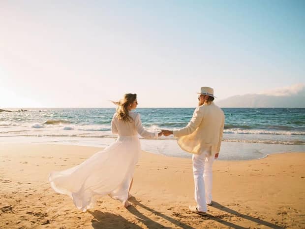 Bride and groom wearing casual white beach wedding attire hold hands on beach