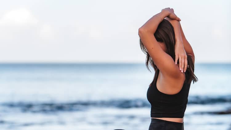 A woman does yoga while sitting on a rock by the ocean