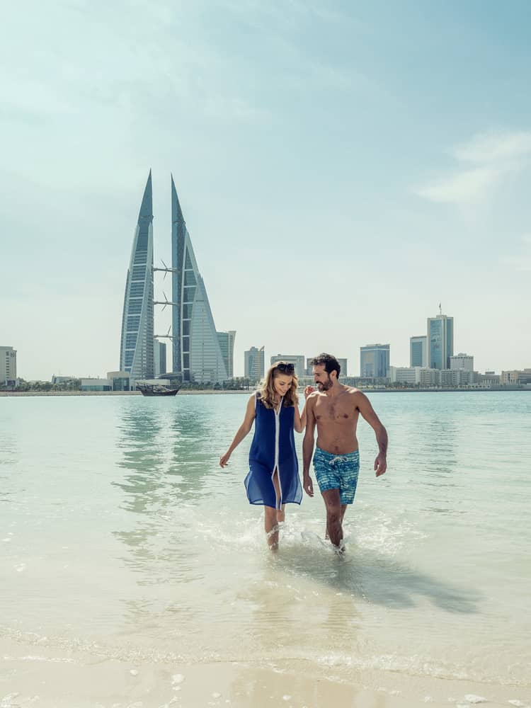 A man and woman run arm in arm through the shallow ocean water with Bahrain Bay skyscrapers in the background
