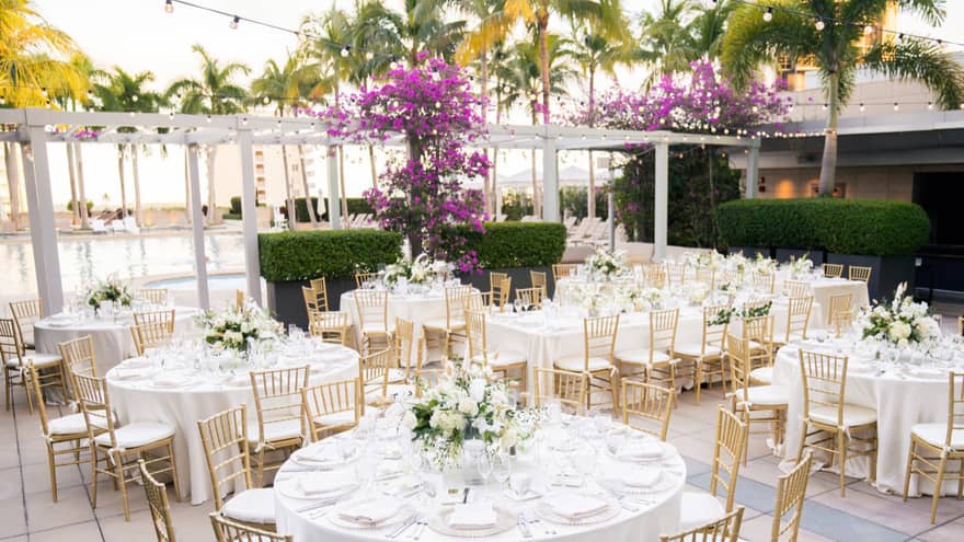 Outdoor venue with tables with bouquets of flowers and palm trees in the background.