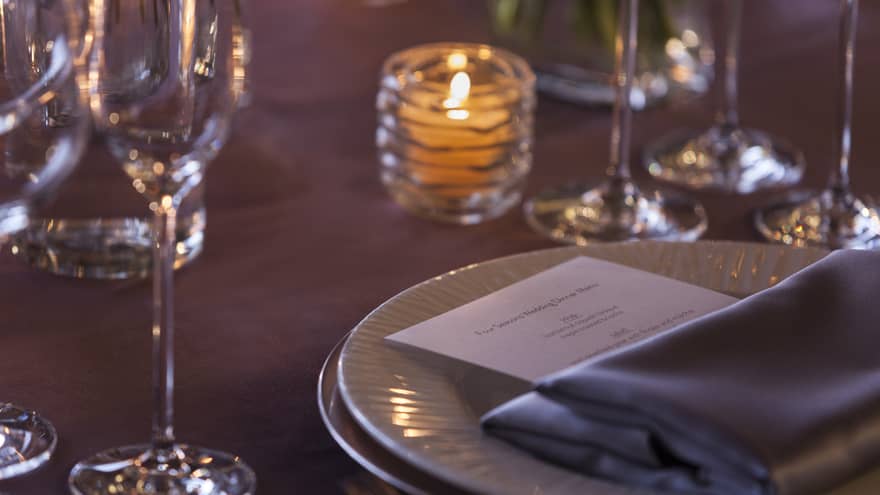 Close up of table with candle, grey napkin and dinner menu on white plate, glasses