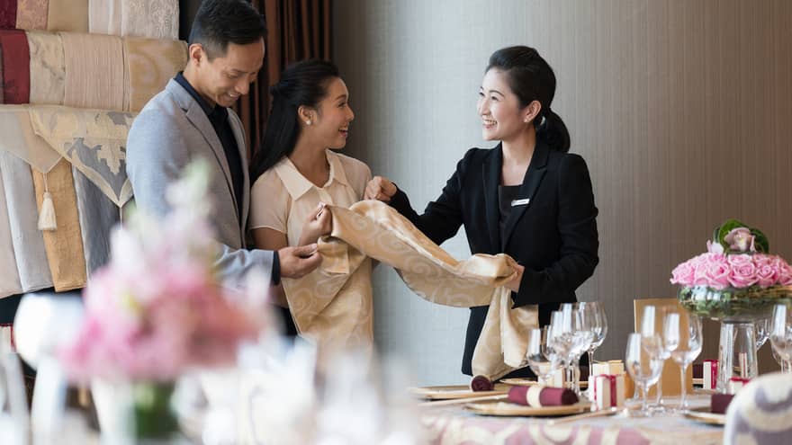 Hotel staff stand by a beautifully set table with flowers and glassware, discussing banquet linen. One explains to others, who smile and engage in conversation