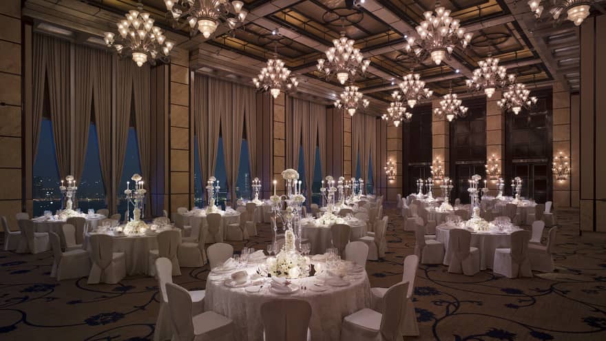 Dimly-lit ballroom with harbor view, round tables with white tablecloths, crystal candelabras, white chairs and floor-to-ceiling windows draped with curtains.