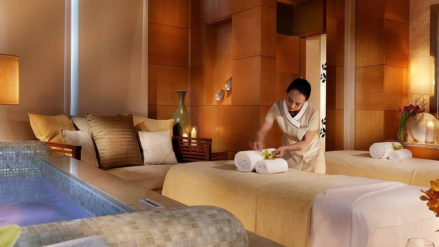 Spa staff sets rolled towels on massage bed near small sofa, tile bathtub in Couples Suite