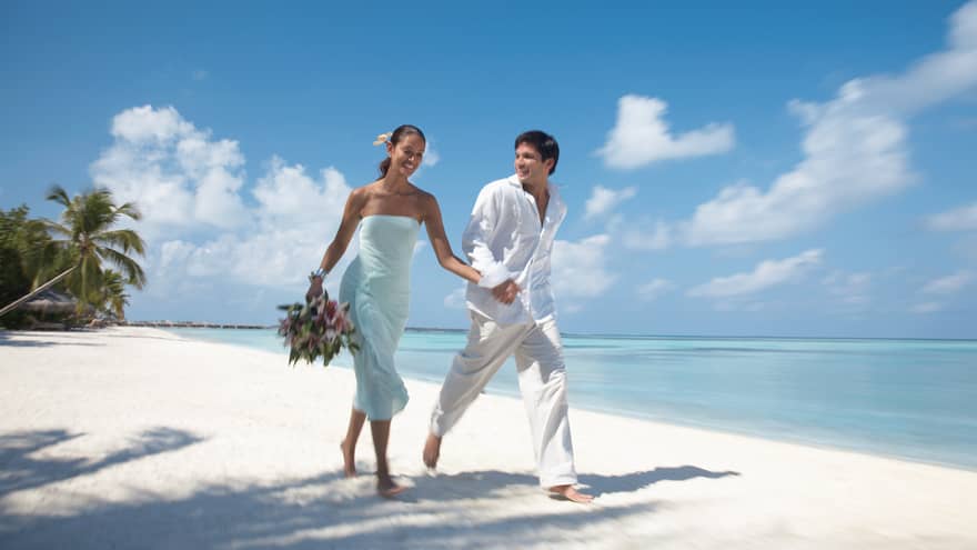 Bride and groom wearing white beach attire walk along sand beach