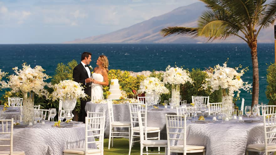 Bride and groom by wedding cake, toast with Champagne near outdoor reception banquet tables
