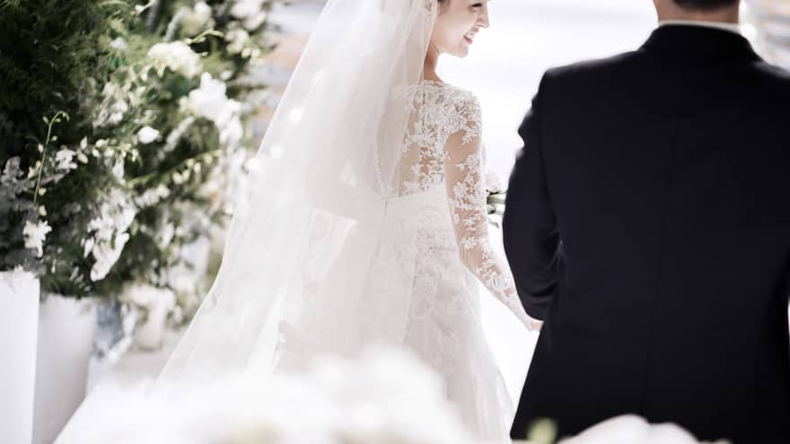 A bride in a white, lace, long sleeve gown smiles as she walks down the aisle with her groom. They are surrounded by artfully arranged white flowers.