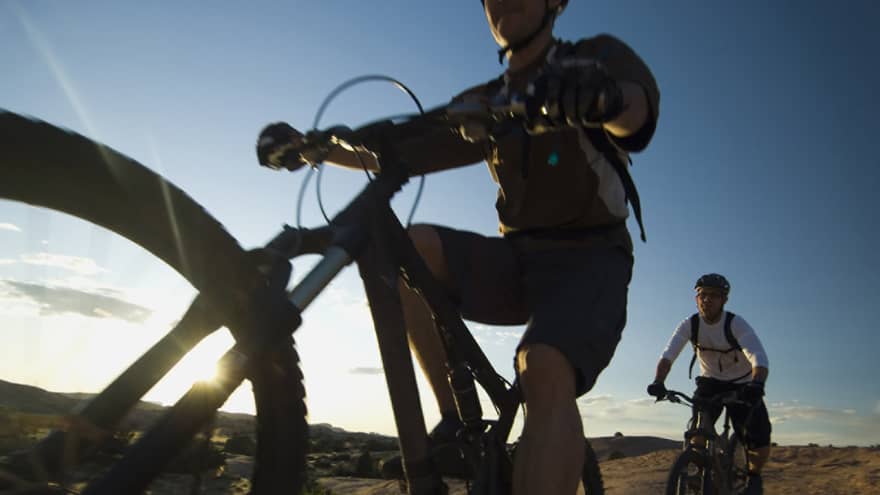 Close-up of cyclist in gear and helmet looking up from bike, second cyclist nearby as sun shines from behind